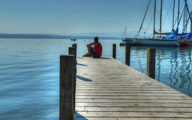 Person sitzt auf einem Holzsteg am Wasser, Boote im Hintergrund.