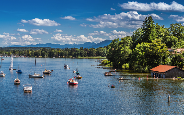 Segelboote auf einem See mit bewaldeten Ufern und Bergen im Hintergrund.