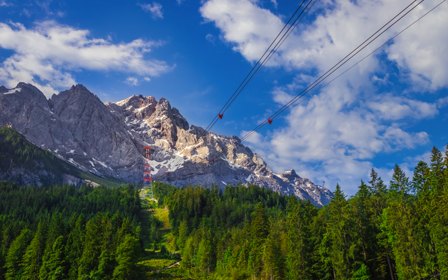 Berglandschaft mit Seilbahn, Wald im Vordergrund, blauer Himmel mit Wolken.
