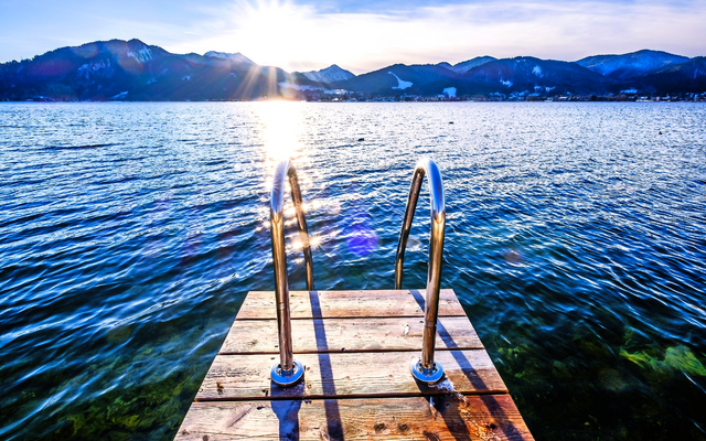 Steg mit Geländer führt ins blaue Wasser eines Sees, Berge im Hintergrund, Sonnenuntergang.