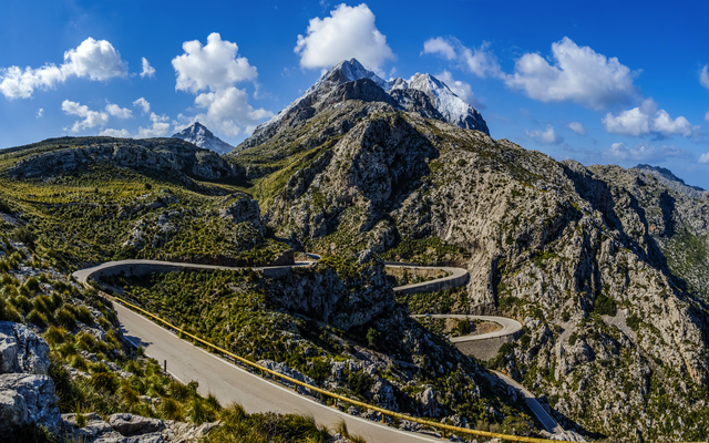 Serpentinenstraße in bergiger Landschaft mit blauem Himmel und Wolken.