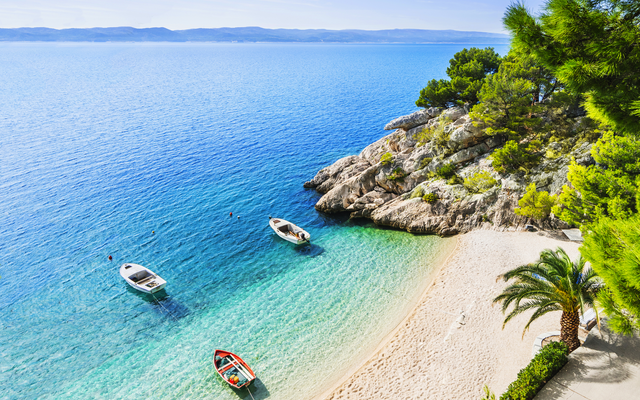 Strand mit klarblauem Wasser, Booten, Felsen, Bäumen und weißem Sand