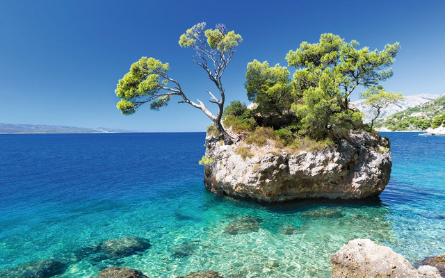 Kleiner Felsen mit Bäumen im klaren blauen Meer vor Küstenlandschaft.