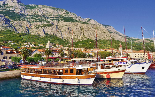 Boote liegen im Hafen vor einer Stadt am Fuße eines großen Gebirges mit grüner Vegetation.