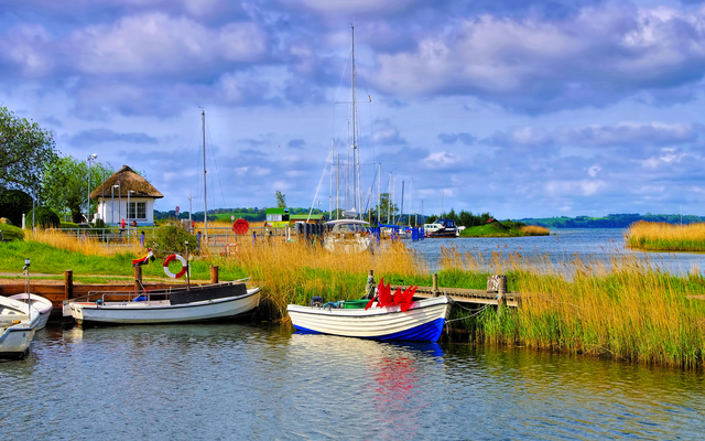 Boote an einem Seeufer mit Schilf und einem kleinen Haus im Hintergrund.