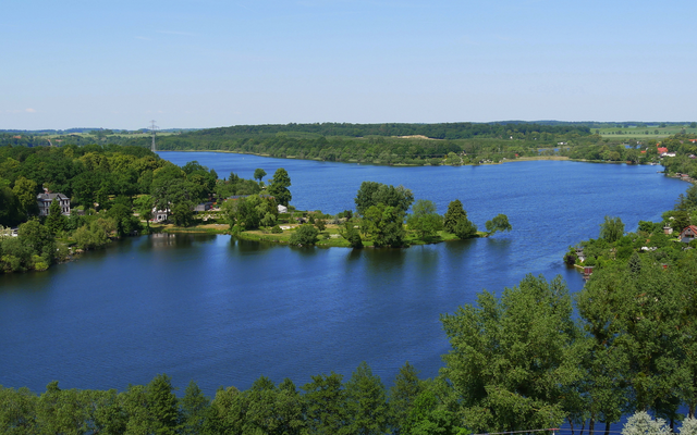 Blick auf einen ruhigen Fluss mit Bäumen und Hügeln im Hintergrund.