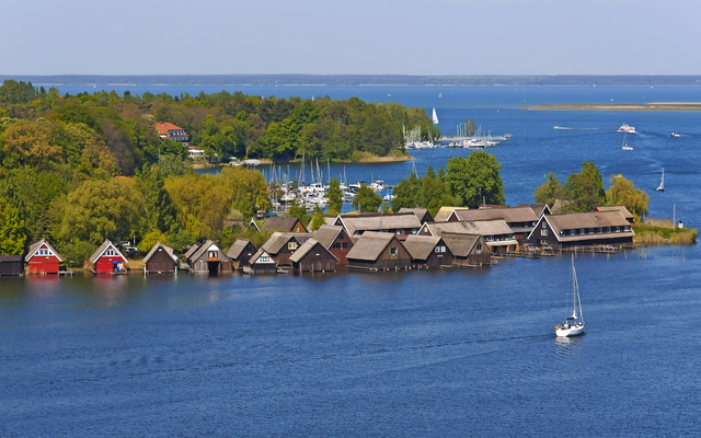 Bootshäuser am Ufer eines Sees mit umliegenden grünen Bäumen und Segelbooten im Wasser.