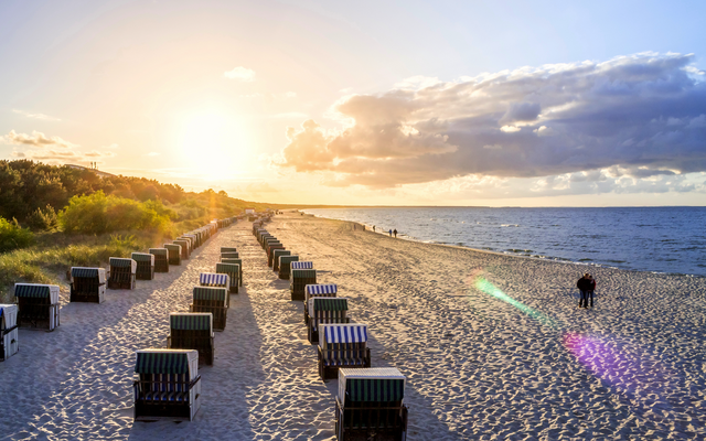 Strand mit Strandkörben bei Sonnenuntergang, Meer und Spaziergängern im Hintergrund.