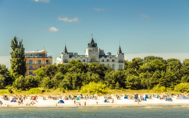 Strand mit Badegästen, dahinter ein Gebäude mit Türmen und Bäumen.