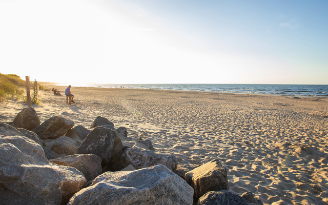 Sonniger Strand mit Meer und Felsen im Vordergrund.