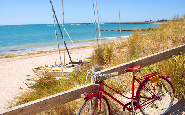 Rotes Fahrrad an einem Holzsteg am Strand mit Segelbooten im Hintergrund.