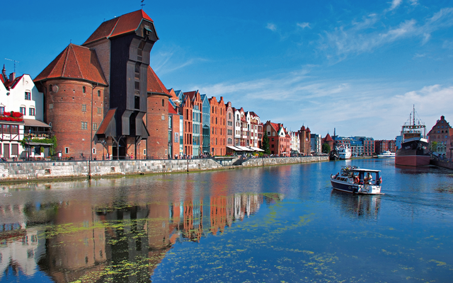 Hafenansicht mit historischem Krantor und Boot auf dem Wasser unter blauem Himmel.