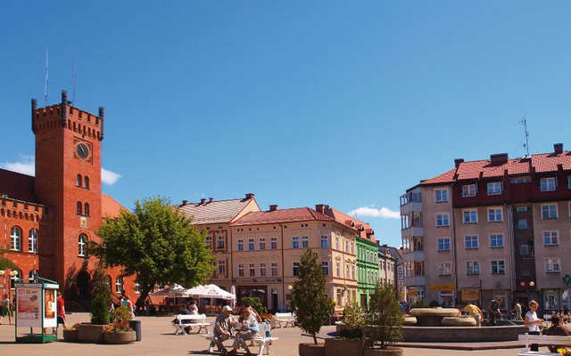 Historischer Platz mit rotem Backsteinturm und umliegenden Gebäuden bei blauem Himmel.