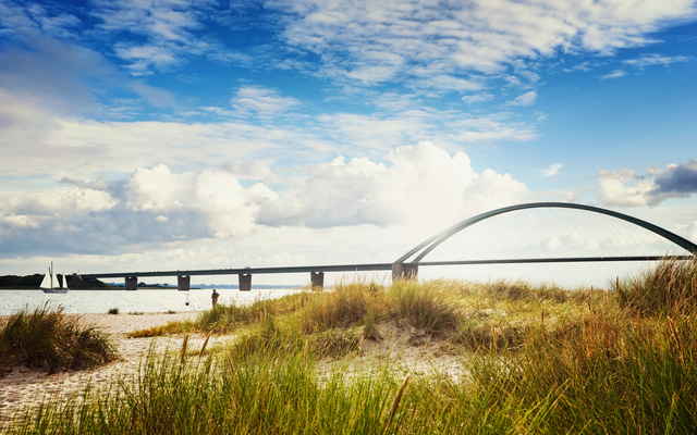 Sandstrand mit Dünengras, im Hintergrund eine Brücke über das Meer bei sonnigem Wetter.