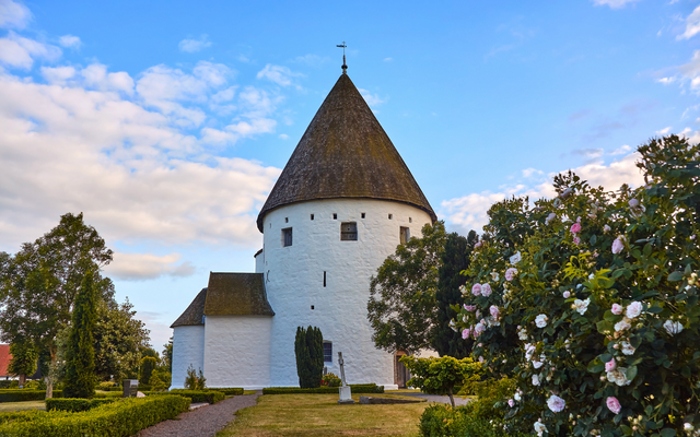 Runde Kirche mit Spitzdach, umgeben von Bäumen und blühenden Sträuchern.