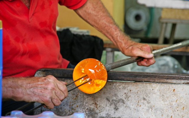 Ein Glasbläser in rotem Hemd formt glühendes, orangenes Glas mit Werkzeugen.