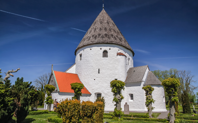 Rundkirche mit weißem Putz und spitzem Dach umgeben von Bäumen und Sträuchern.