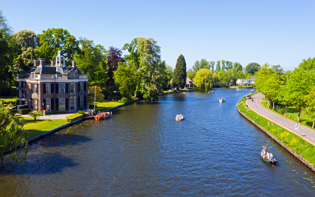 Fluss mit historischen Häusern und Booten, umgeben von grüner Landschaft.