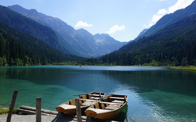 Zwei Ruderboote an einem klaren Bergsee, umgeben von bewaldeten Bergen.
