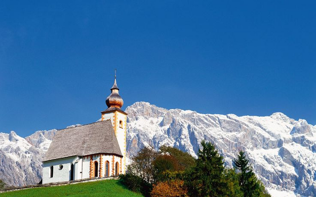Bergkirche St. Nikolaus vor schneebedeckten Alpen und blauem Himmel.
