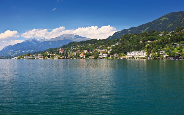 Landschaft am Millstätter See mit Bergen, Häusern und grünem Ufer unter einem blauen Himmel.