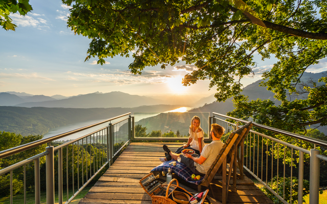 Zwei Personen picknicken auf einem Balkon mit Blick auf den Millstätter See bei Sonnenuntergang.