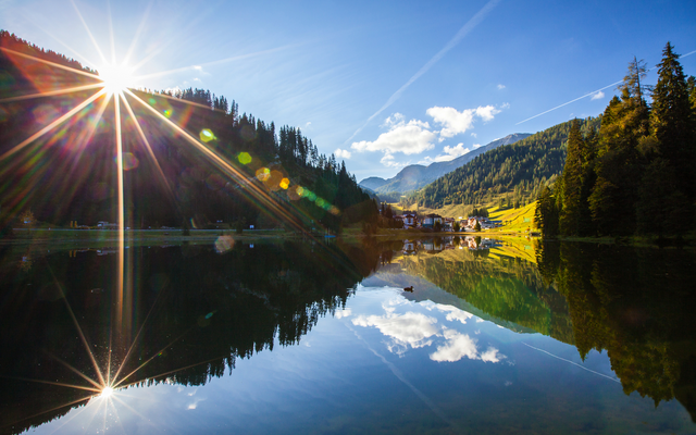 Bergsee mit Bäumen und Sonnenstrahlen im Hintergrund.