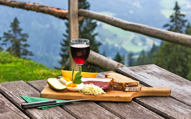 Brett mit Brot, Käse, Fleisch und Wein auf Holztisch, Bergblick im Hintergrund.