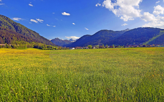 Grüne Wiese vor bergiger Landschaft unter blauem Himmel mit Wolken.