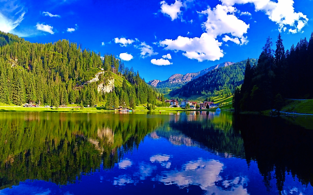 Bergsee mit Wald und klarer Spiegelung im Wasser unter blauem Himmel mit Wolken.