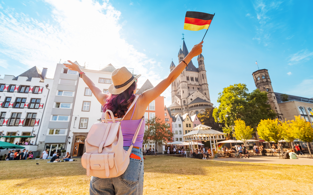 Frau mit Rucksack hält deutsche Flagge in einer Altstadt mit historischer Kirche.