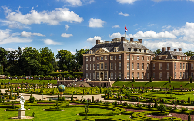 Historisches Gebäude mit angelegtem Garten und blauen Himmel im Hintergrund.