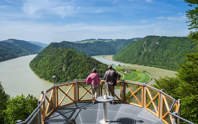 Aussichtsplattform mit Blick auf eine Flusslandschaft und bewaldete Hügel im Sonnenlicht.