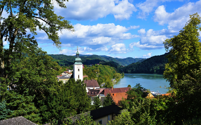 Landschaft mit Fluss, einem Kirchturm, Häusern und bewaldeten Hügeln im Hintergrund.