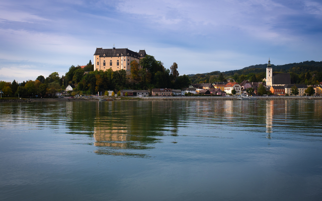 Flusslandschaft mit Schloss und Kirche im Hintergrund, sanfte Hügel und bewölkter Himmel.