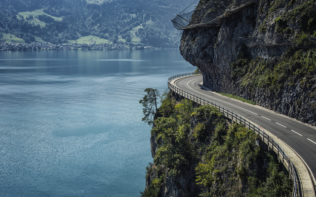 Straße entlang eines felsigen Abgrunds mit Blick auf einen See und bewaldete Hügel.