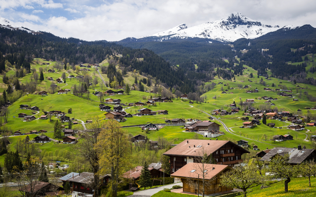 Bergdorf mit traditionellen Holzhäusern, grünen Wiesen und schneebedeckten Bergen im Hintergrund.