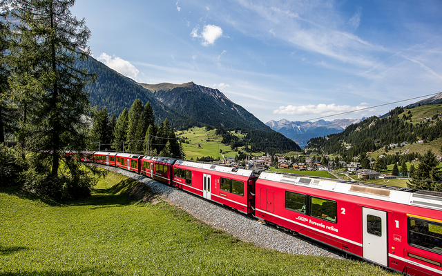 Roter Zug fährt durch grüne Albulatal-Landschaft mit Bergen im Hintergrund.