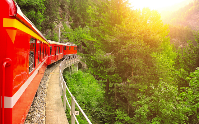 Roter Zug auf einer kurvigen Strecke durch bewaldete Berglandschaft bei Sonnenuntergang.
