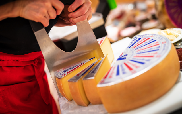 Person schneidet großen Laib Käse mit einem Käsemesser in einem Laden.