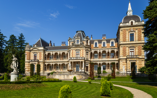 Großes historisches Gebäude mit Garten und Statue im Vordergrund.