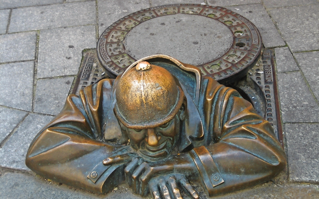 Bronzestatue eines Mannes mit Helm, der aus einem Gully-Deckel auf dem Gehweg schaut.