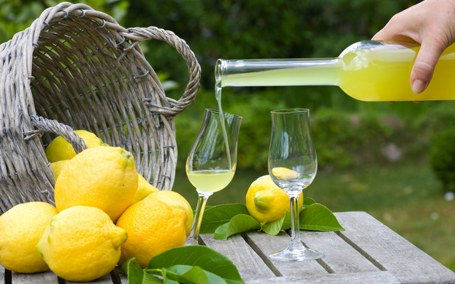 Eine Hand gießt Limoncello in Gläser auf einem Tisch, neben einem Korb voller Zitronen im Freien.