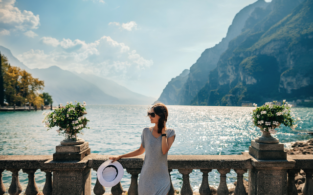 Frau steht an einem Geländer mit Blick auf einen See und Berge im Hintergrund.