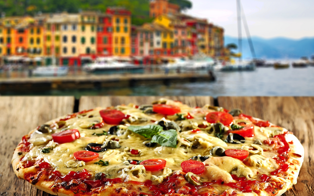 Pizza mit Käse und Gemüse auf Holztisch mit malerischem Küstenhintergrund und bunten Häusern.