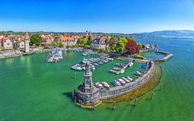 Panorama eines Hafens mit Segelbooten und Löwenstatue, umgeben von Gebäuden und See.