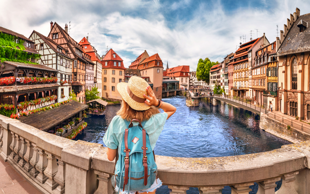 Frau mit Hut und Rucksack schaut auf malerische Fachwerkhäuser an einem Fluss.