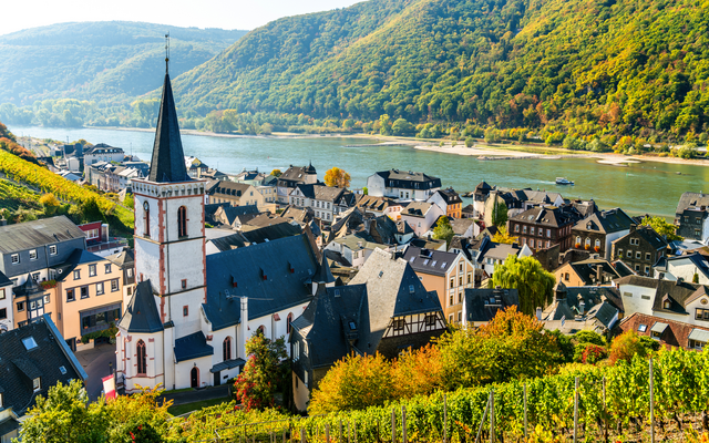 Blick auf ein Dorf mit Kirche am Fluss, umgeben von Weinbergen und bewaldeten Hügeln.