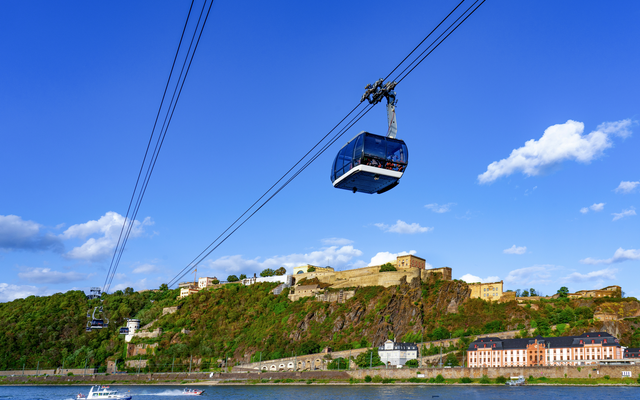 Seilbahn über einem Fluss mit Festung im Hintergrund bei blauem Himmel.