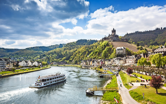 Flusslandschaft mit Boot, Hügel, mittelalterliche Burg und Stadt am Ufer unter blauem Himmel.